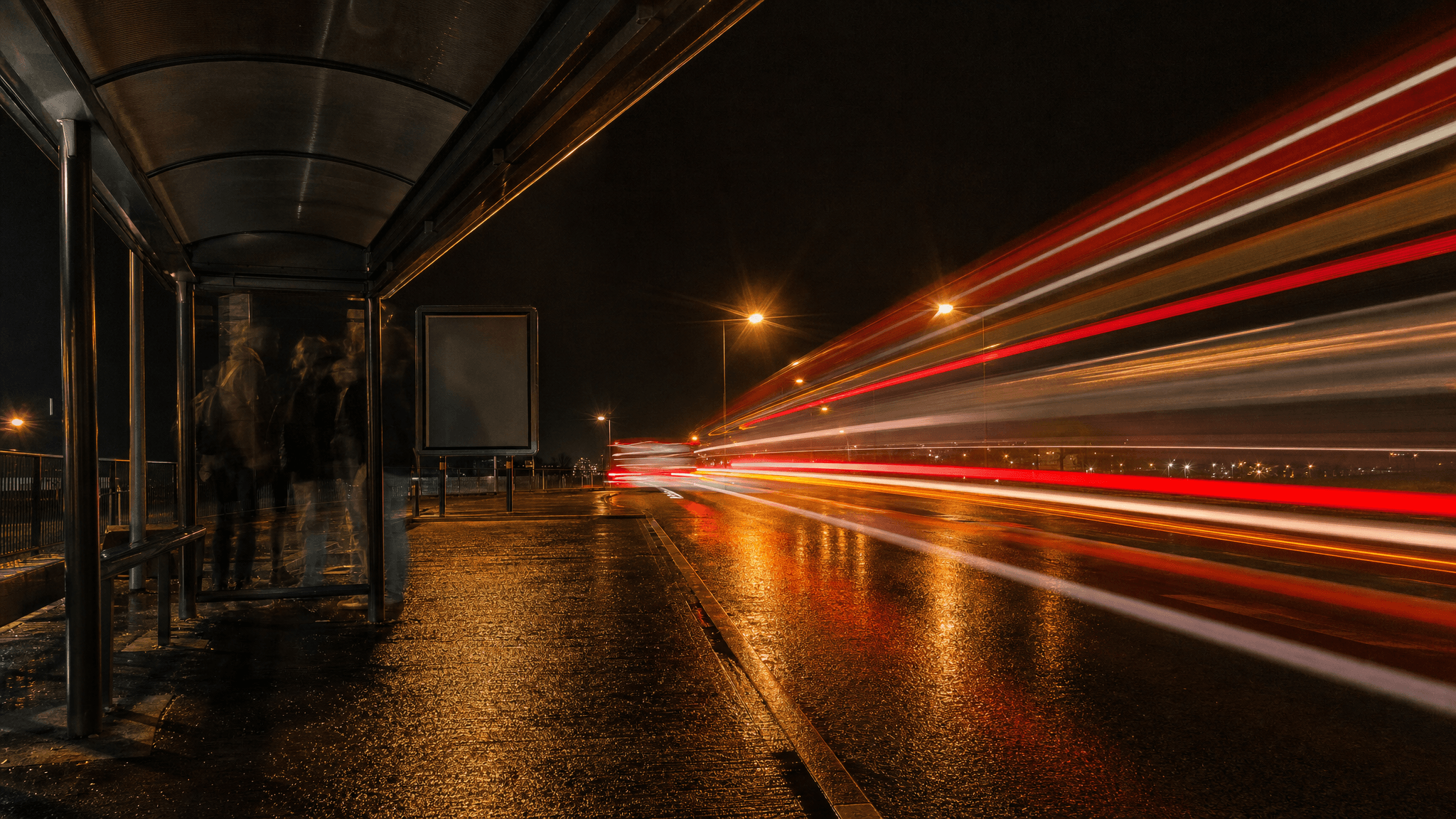 This video captures a dynamic, moody, and somewhat mysterious night-time urban scene. The setting is an outdoor street or highway at night, with a bus stop or shelter on the left side. The activity level is a blend of static (people waiting) and highly dynamic (fast-moving light trails from vehicles). The visual style is realistic, employing long exposure photography techniques to create artistic light streaks. The emotional tone is contemplative, perhaps a little melancholic, with a sense of movement and passage of time. The subjects include blurred, ghostly figures of people waiting at the bus stop, a street lamp, and the vibrant red and white light trails of cars speeding by. The artistic style leans towards photorealism with an artistic long-exposure effect. The color characteristics are dominated by warm tones from the streetlights and vehicle lights, contrasted with the cool, dark night sky and wet asphalt. The color palette primarily features deep blacks, dark grays, and rich oranges and reds, with streaks of white.