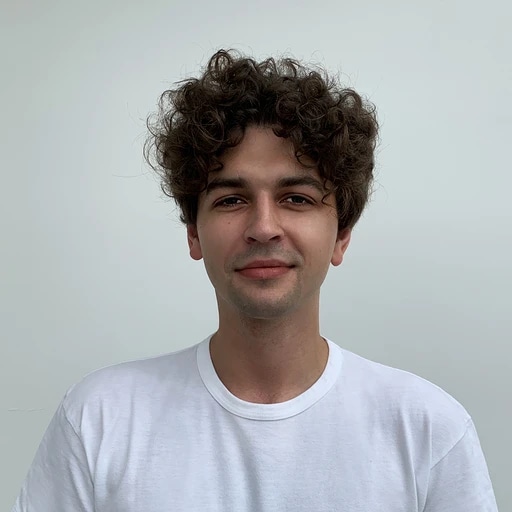 A young man with curly hair wearing a white t-shirt, smiling softly against a plain background.