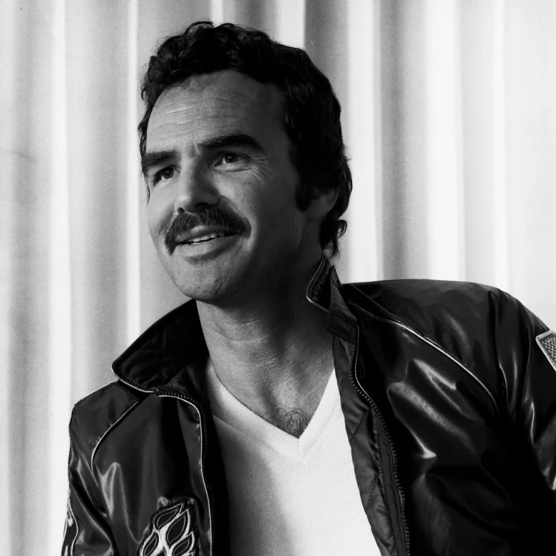 A man with a mustache and curly hair, wearing a leather jacket and a white shirt, looking to the side.