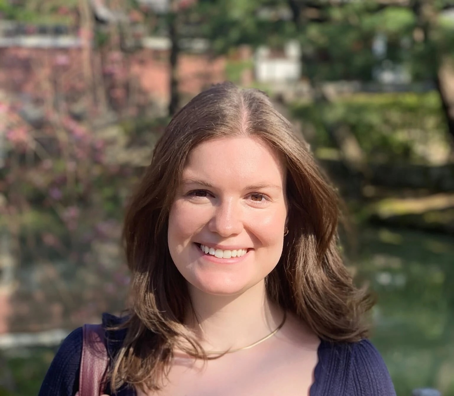 A woman smiling outdoors with trees and a building in the background.